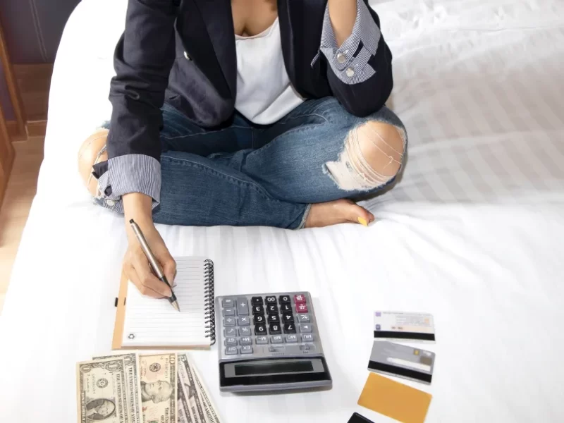 A woman sitting on a bed, using a calculator, pen and money to manage a personal loan with bad credit.