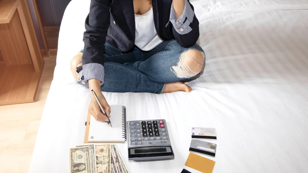 A woman sitting on a bed, using a calculator, pen and money to manage a personal loan with bad credit.