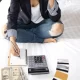 Une femme assise sur un lit, utilisant une calculatrice, un stylo et de l'argent, réfléchissant à un prêt personnel avec un mauvais crédit.