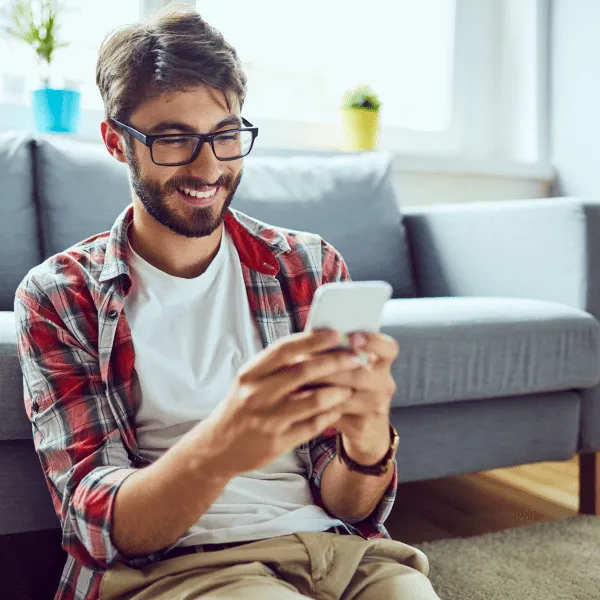 A man in glasses sits on the floor, engrossed in using his phone