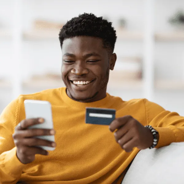 A man happily holds a credit card while attentively looking at his phone, displaying a cheerful expression