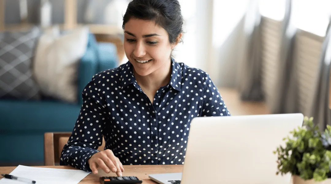 A woman sitting at a table, working on a laptop and using a calculator to perform calculations.