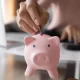 Une femme insérant des pièces dans une tirelire en forme de cochon, symbolisant l'épargne et le refinancement.